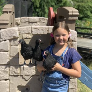 Smiling Georgia is standing in front of a faux brick play structure. Two black hens are on the wall. She is holding a third chicken in her hands.