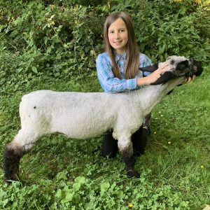 Annalise with her lamb in a green field with greenery behind her.