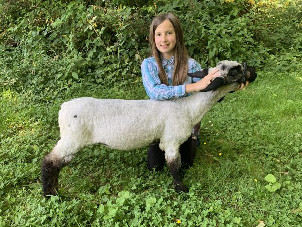 Annalise with her lamb in a green field with greenery behind her.