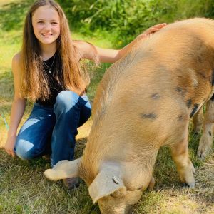 Smiling Emme kneeling next to her hog, Wilma, who is eating grass.