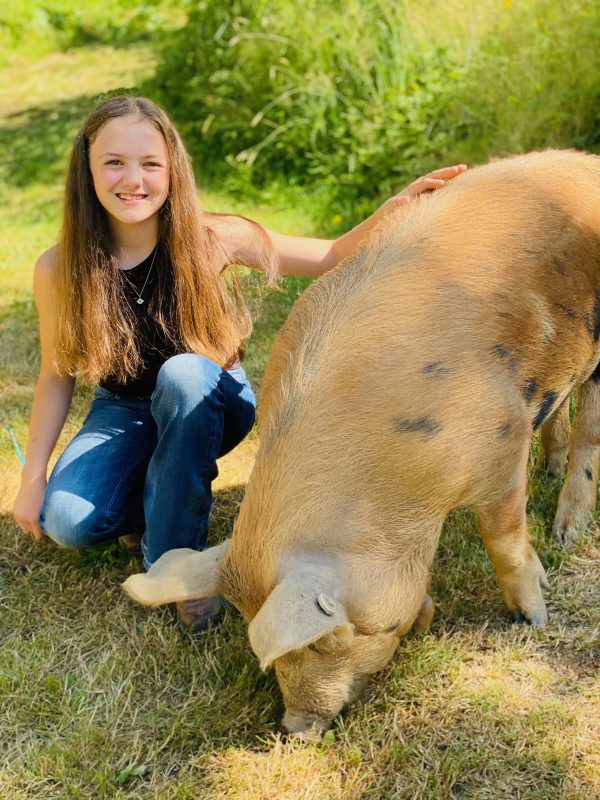 Smiling Emme kneeling next to her hog, Wilma, who is eating grass.